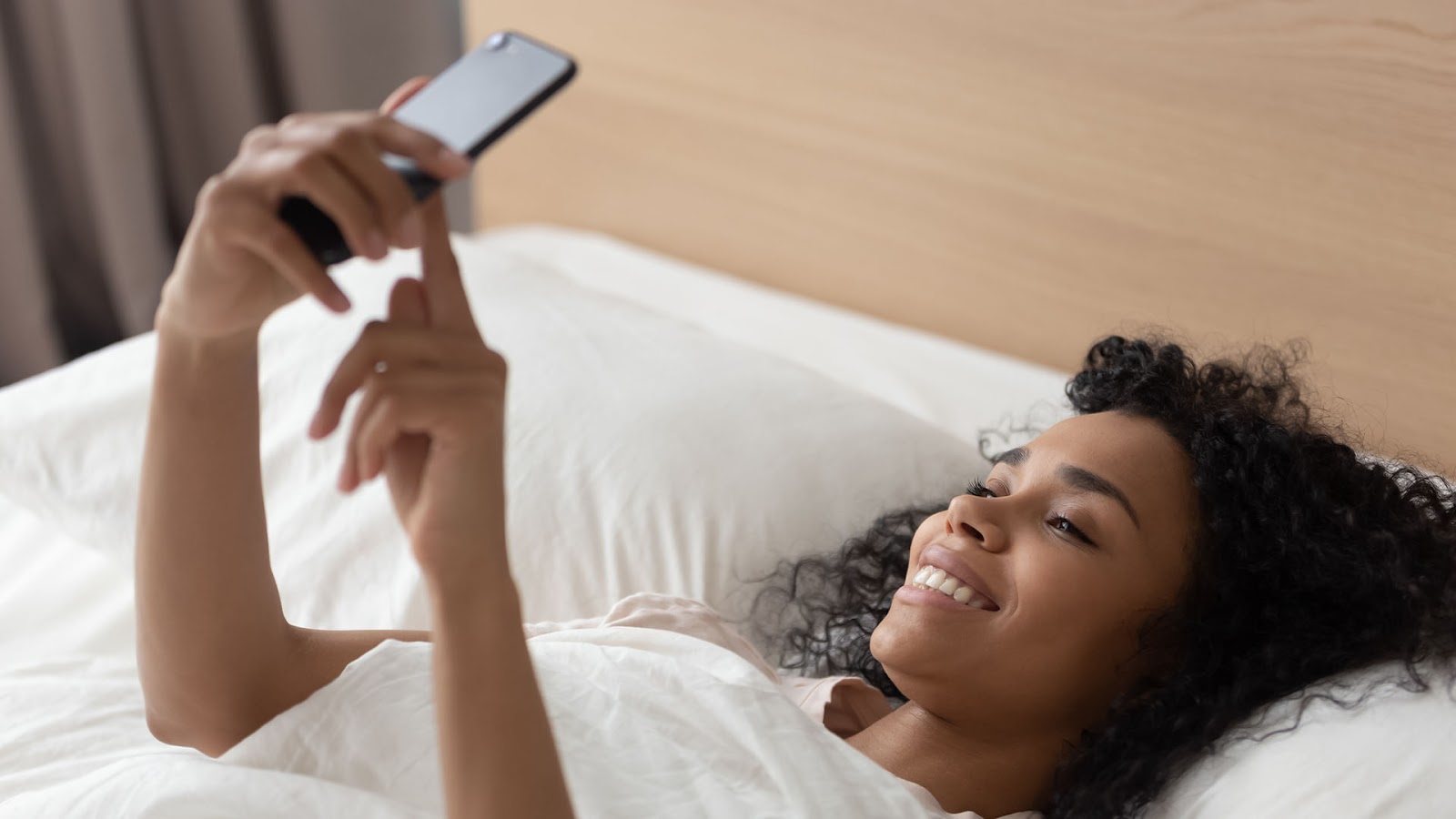 Brunette woman with curly hair lying in bed of white sheets, and smiling while looking at her phone