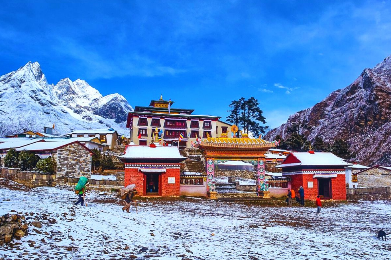 Snow-covered landscape featuring a traditional Sherpa monastery with colorful prayer flags, surrounded by mountains, and trekkers exploring the area.