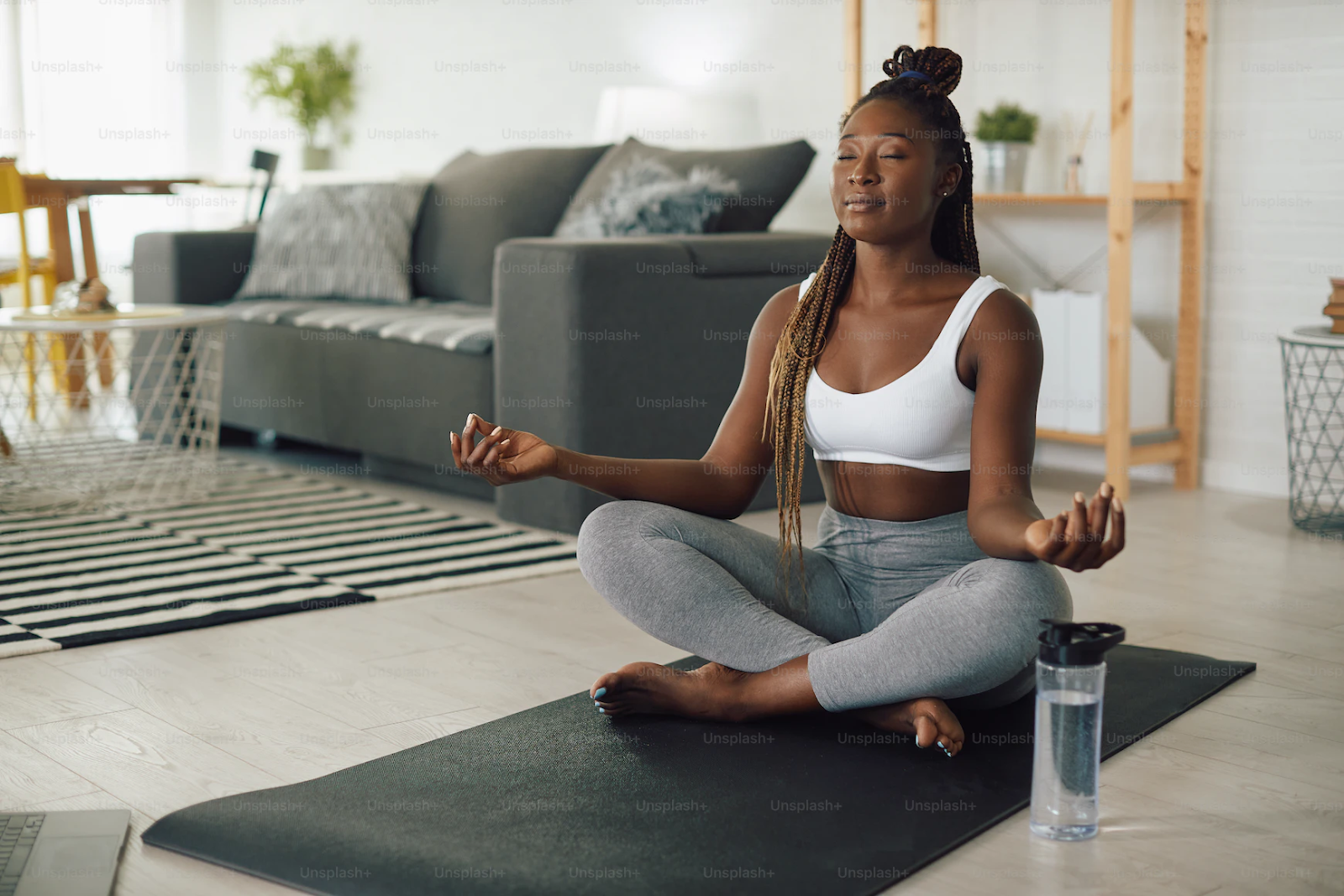 A black woman with braids, wearing a white sports bra and gray leggings, meditates on a yoga mat - How to Navigate Social Anxiety in College