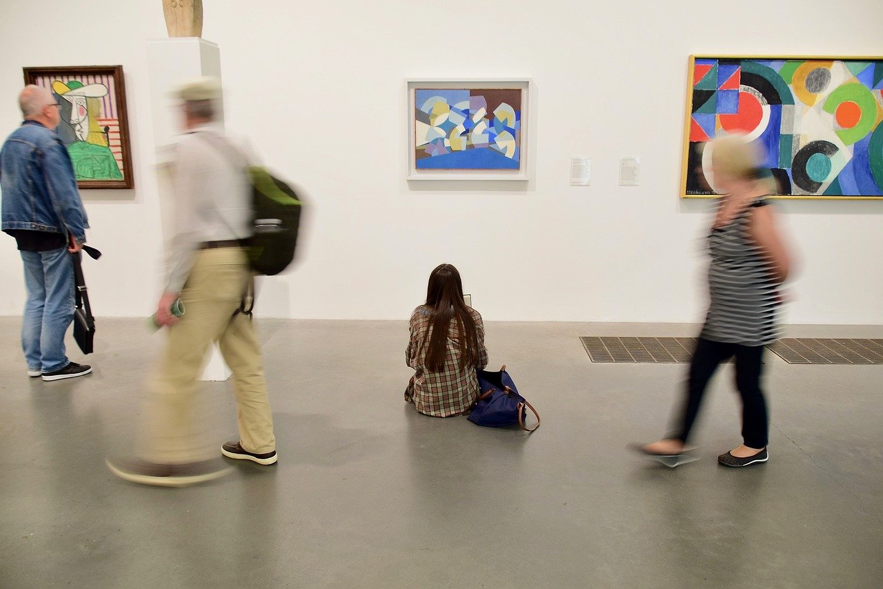 a gallery visitor in a flannel coat sitting on the floor of an art gallery and looking up at several colorful gallery art prints