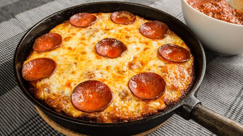 A freshly baked deep-dish pepperoni pizza in a cast iron skillet, with bubbly melted cheese and crispy-edged pepperoni, accompanied by a bowl of marinara sauce on the side.