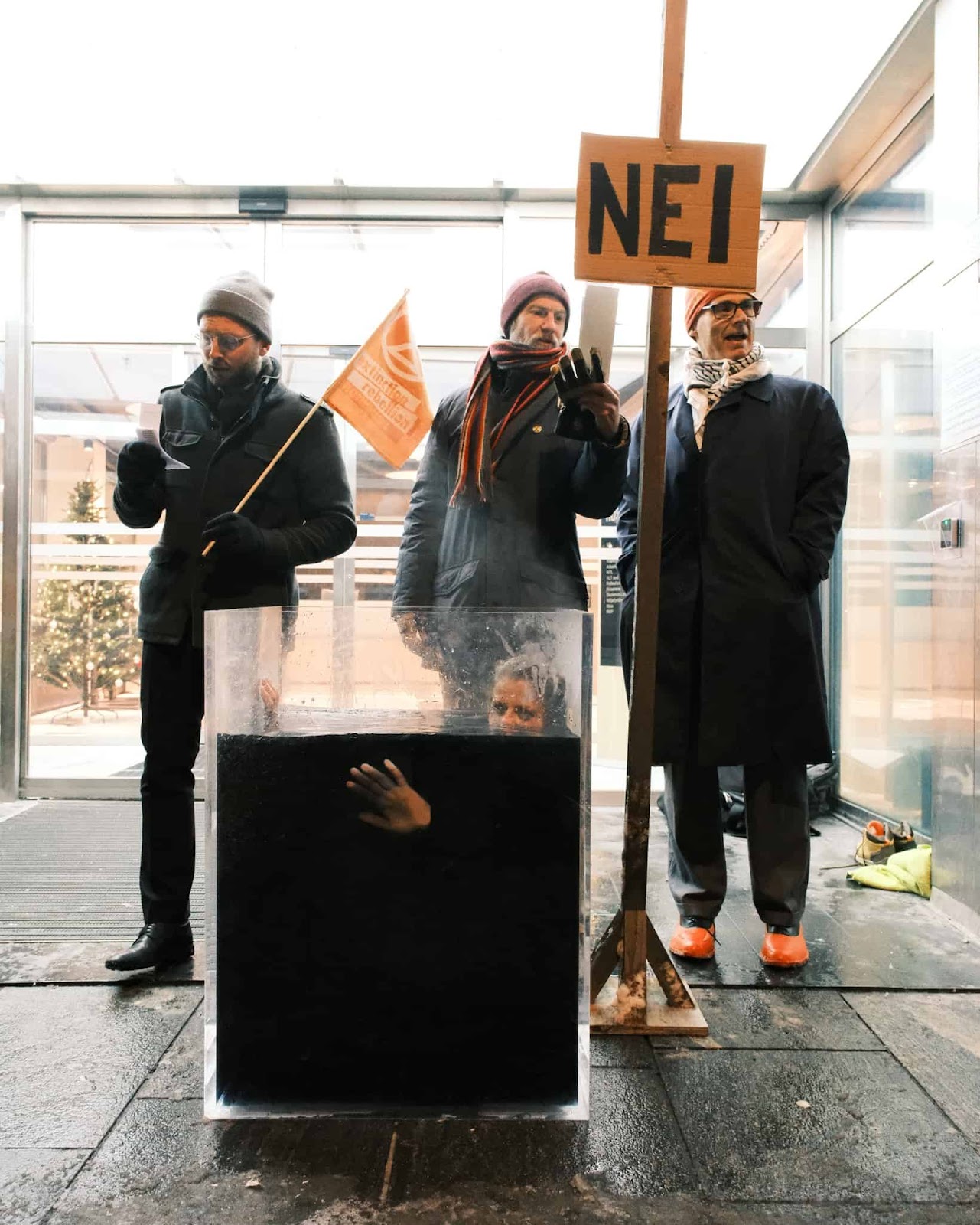 Rebels stand outside a workplace entrance with a sign that says NEI (No) and a rebel sat submerged in a glass tank of black liquid