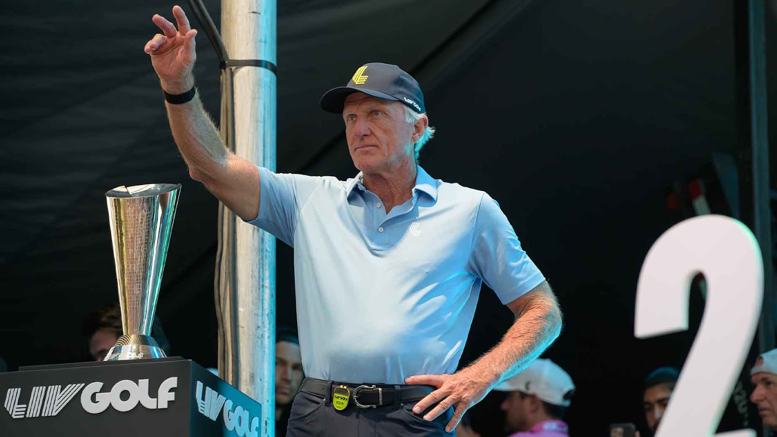 greg norman points in blue shirt from stage at LIV Golf team championships in Miami.