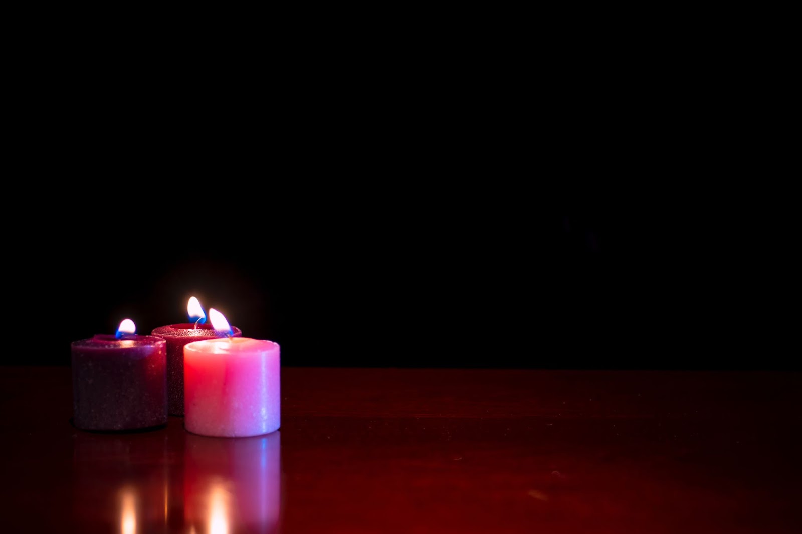 Two purple and one pink candles burning on a hardwood floor with a black background.