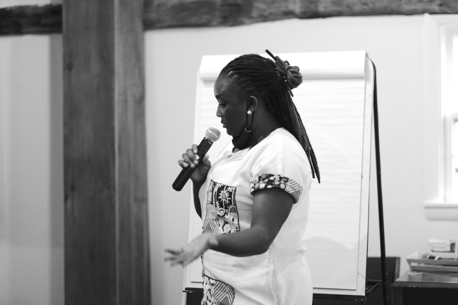 A Ugandan organizer, Peruth, Nabirye in a white linen tunic with colorful accents speaks into a mic in front of a flipchart