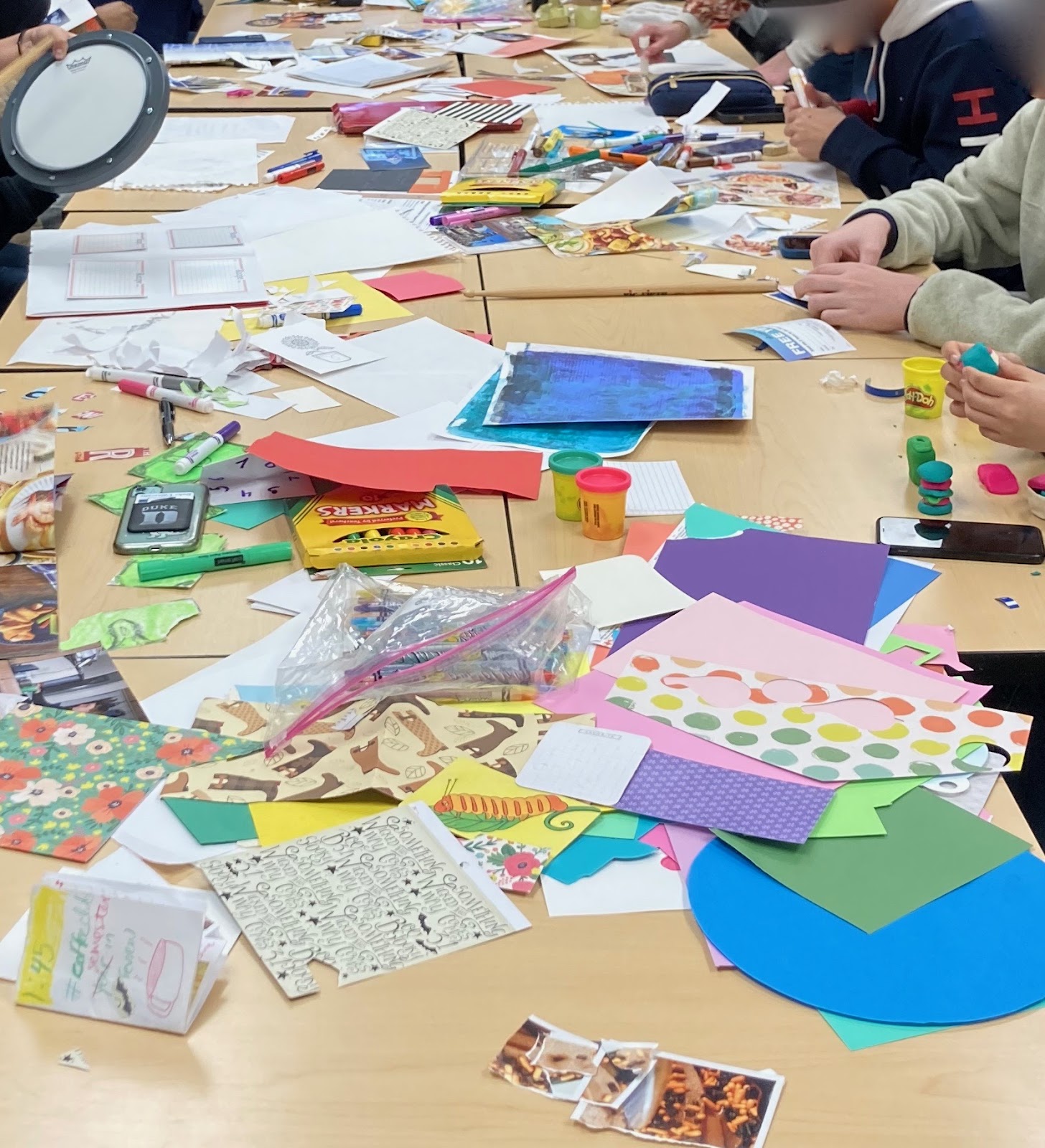An image of a table covered in craft supplies with a few hands visible showing people creating artwork. 