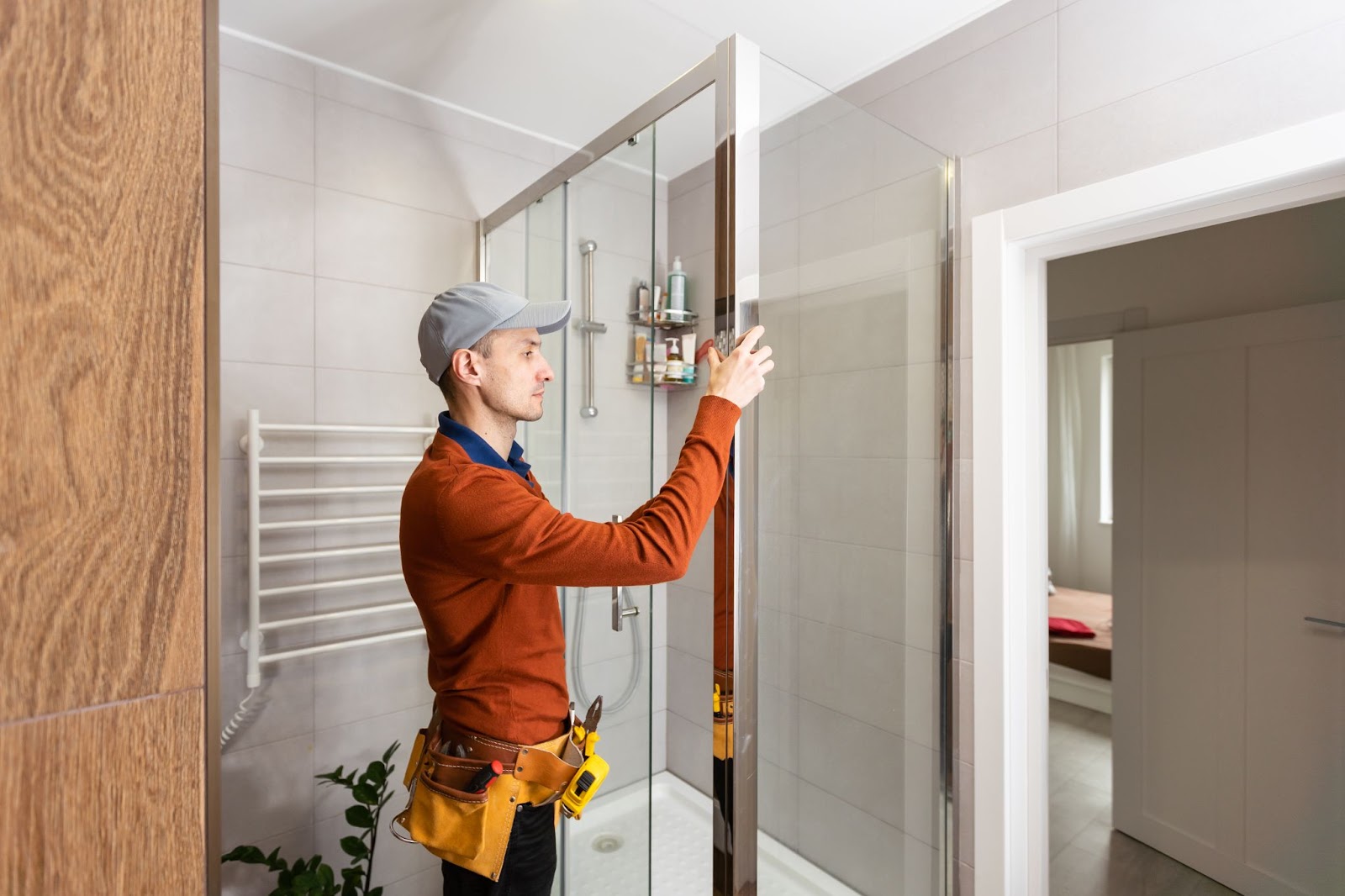 A professional handyman wearing a gray cap and rust-colored uniform with a toolset around his waist works on a replacement shower; a plant and towel rack are visible in the background.