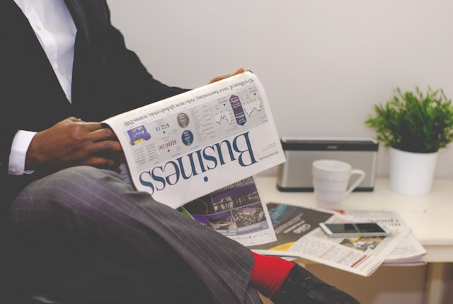 A person in a suit sitting in an office chair reading a business newspaper.