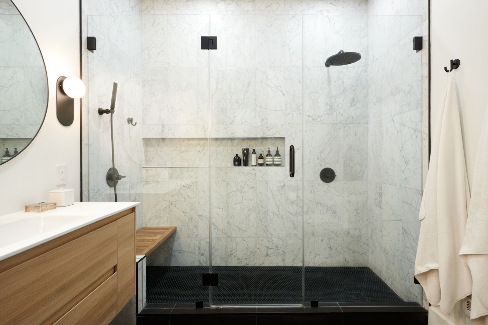 Large contemporary walk in shower with shower bench, niche for storage, matte black fixtures, large format white marbled wall tile, and black floor tile