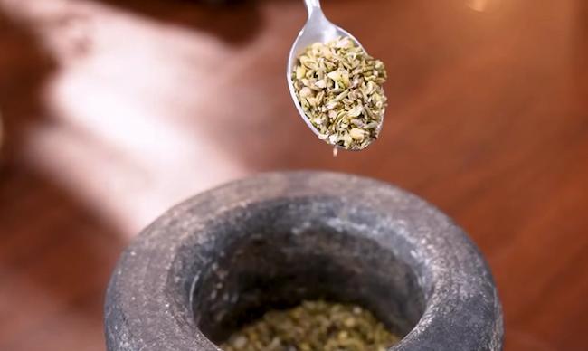 Coarse spice mix of fennel seeds, coriander seeds, and black pepper being pounded in a mortar and pestle