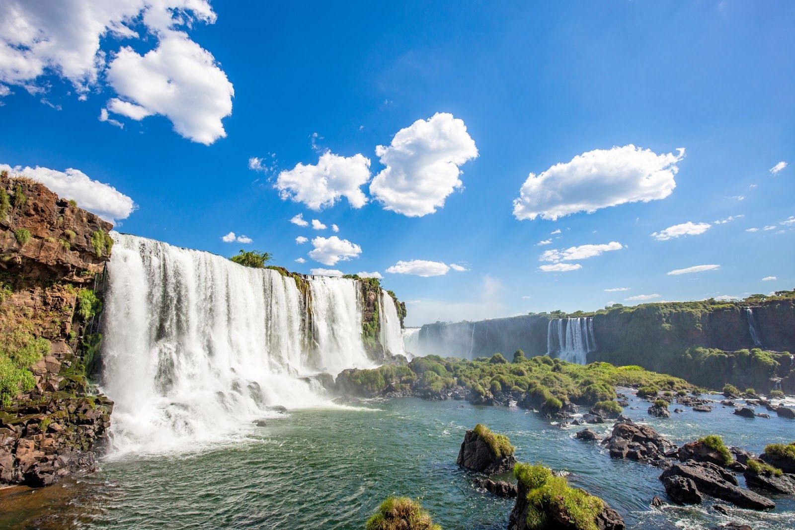 Quedas d’água do Parque Nacional do Iguaçu. Elas caem de paredões cobertos por vegetação rasteira e verde. Ao fundo, o céu azul aparece com poucas nuvens.
