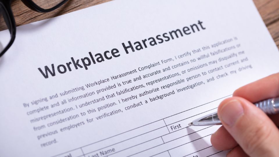 Close-up of a 'Workplace Harassment' complaint form with glasses resting on it and a hand holding a pen, ready to fill in the details.