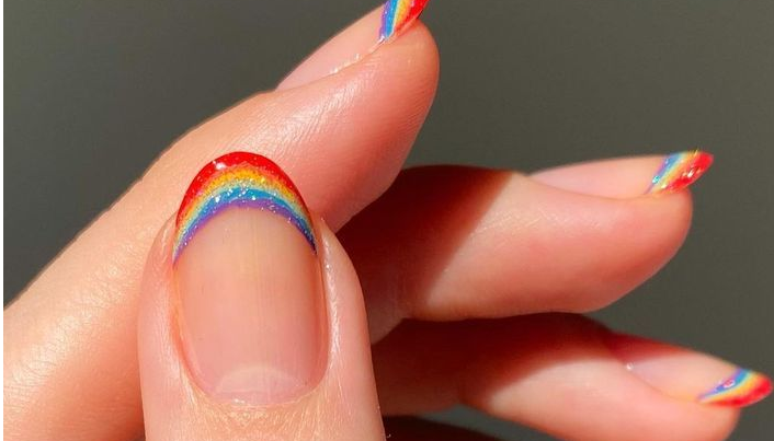 Mão feminina com unhas arredondadas pintadas com uma francesinha formando um arco-íris na ponta dos dedos.