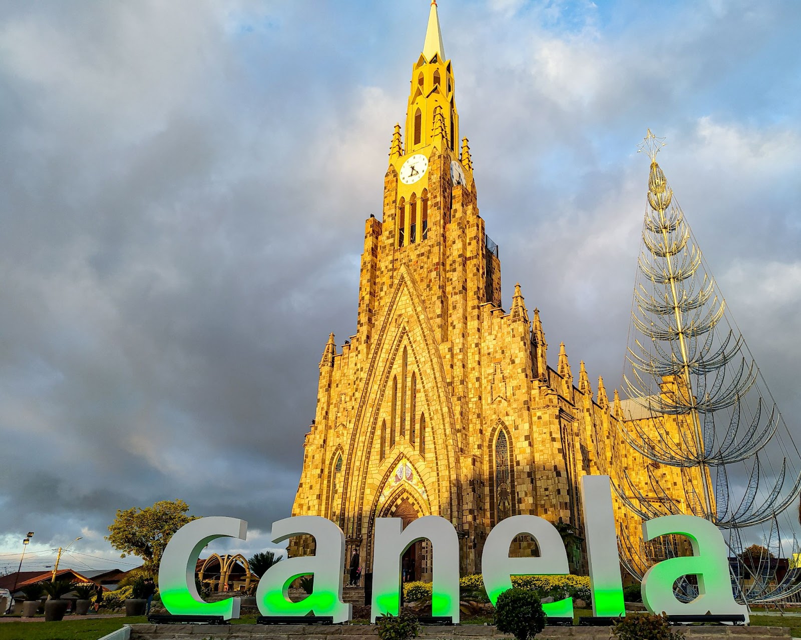 Catedral de Pedra iluminada pela luz dourada do entardecer, com o letreiro escrito “Canela” iluminado por luzes verdes.