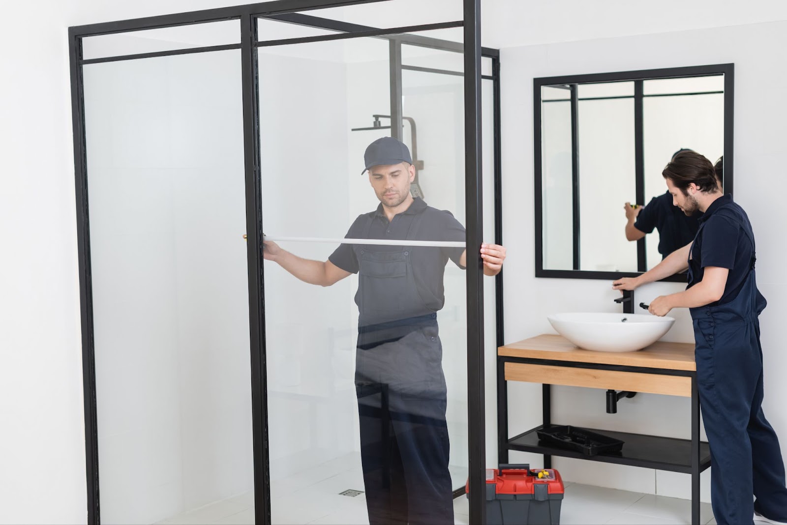 A foreman measures the shower cabin in preparation for a replacement shower door while a plumber works on the sink. Both are wearing black uniforms.