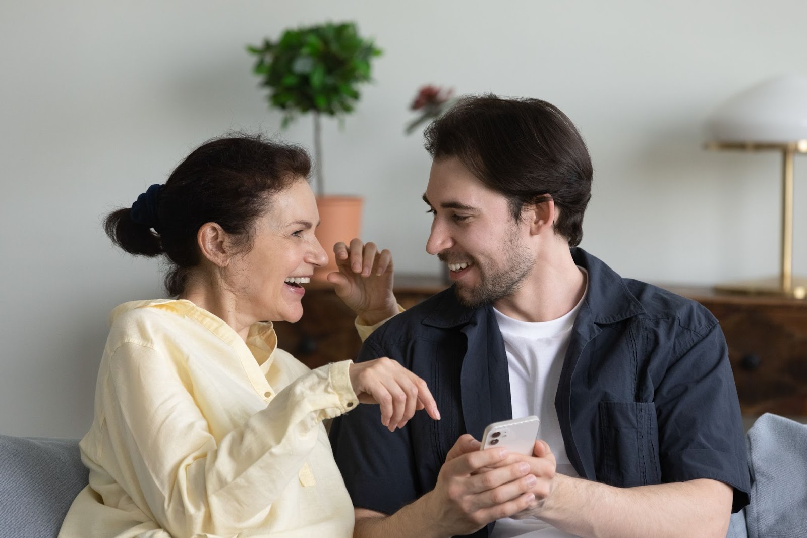 Um jovem e sua mãe sentados lado a lado em um sofá. Os dois sorriem e olham para a tela de um celular.