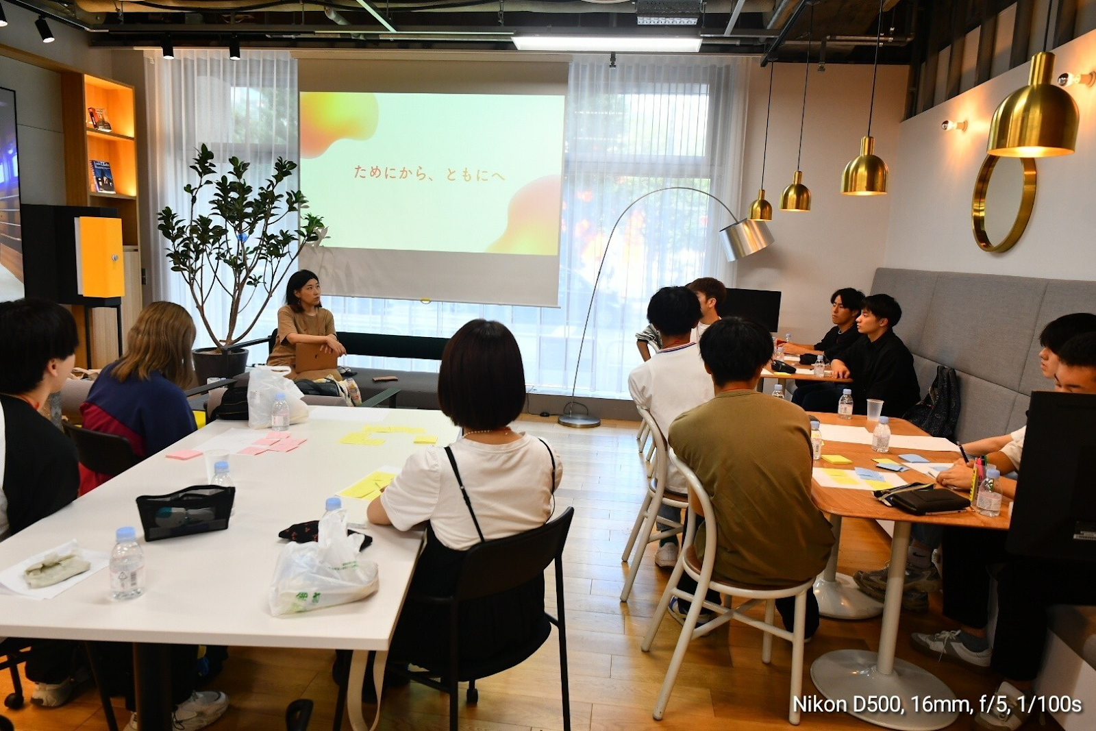 学生さんに３テーブル（１テーブルあたり３〜４人）に分かれて座ってもらいました。中央のスクリーン前のソファーに山田が座って学生さんに向けて何かしらを話しています。
受講生の学生さんは11名写っています。