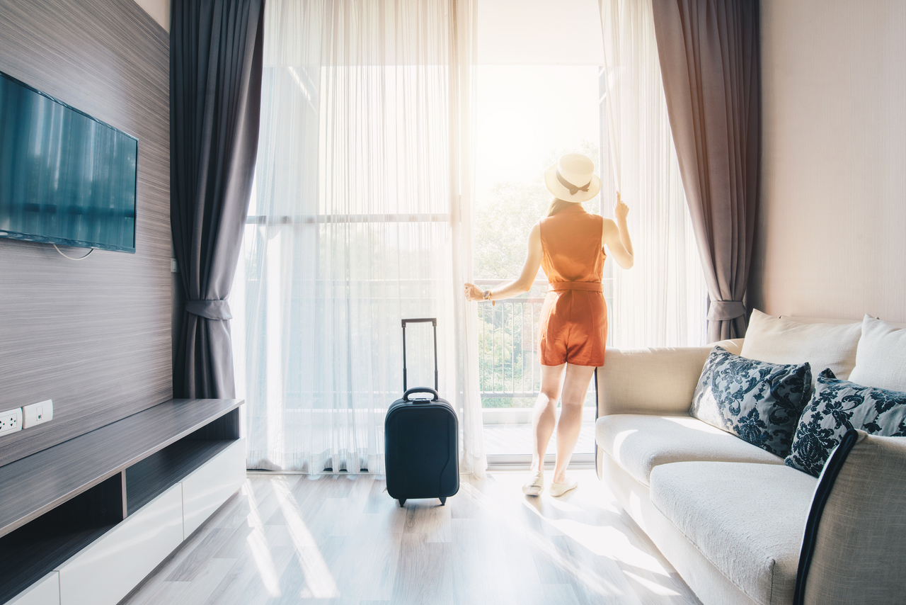 Eine Frau mit Koffer steht vor einem Fenster in einem Hotelzimmer.