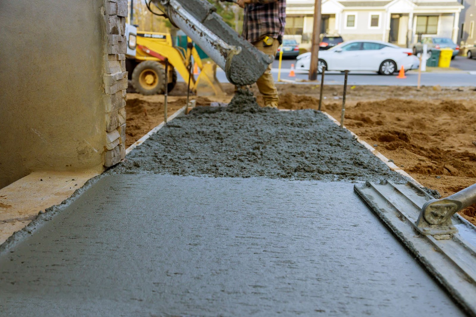A close-up of a construction worker pouring reinforced concrete for the garage and sidewalk of a new home, with a white car and other vehicles parked in soft focus across the street.