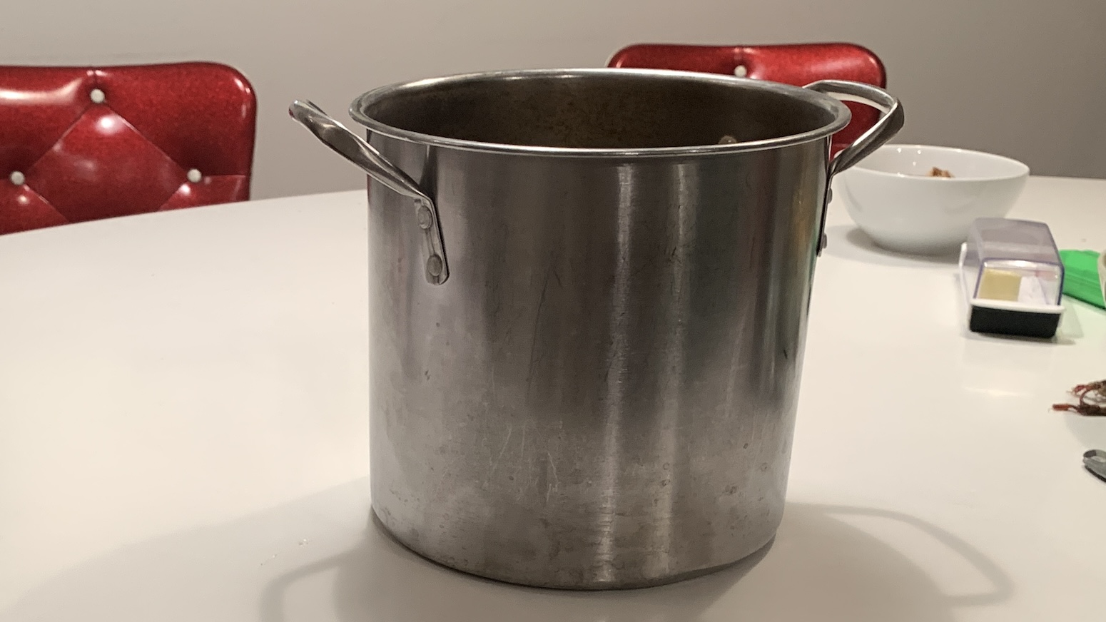 A battered steel pasta pot sits on a white tabletop with shiny red chair-backs showing in the background. A butter dish and bowl are to the right. 
