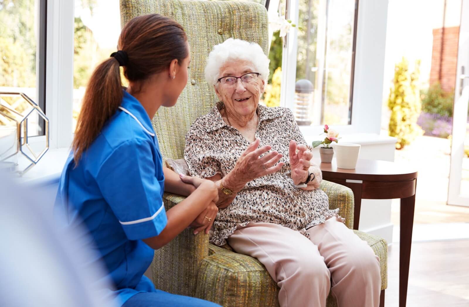 An older adult woman in a memory care facility sitting on a chair smiling and having a conversation with a nurse.