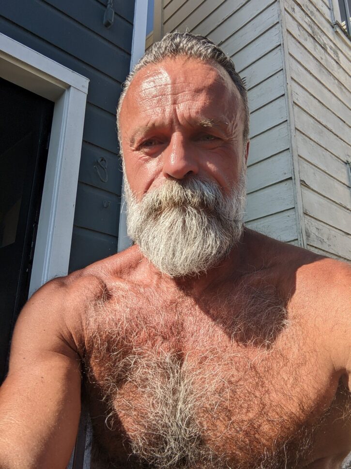 Daddy John shirtless outside on a sunny day taking an iphone selfie on the patio