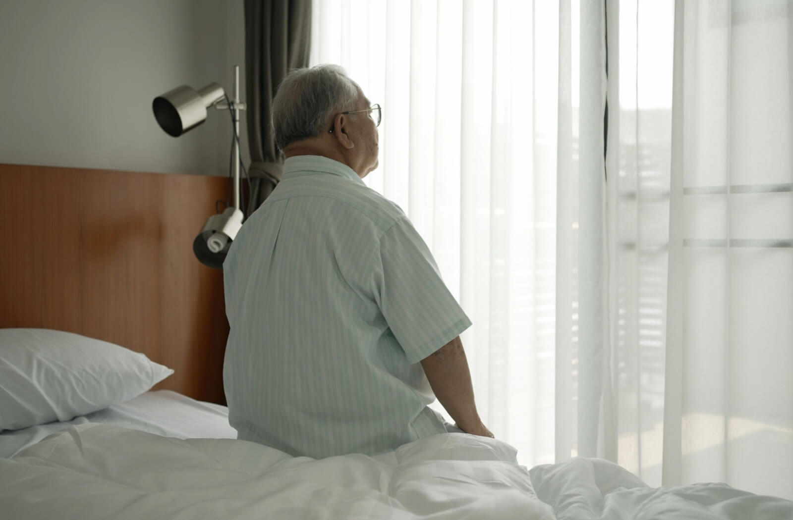 An older adult man sitting alone in his bedroom while  he is spacing out.