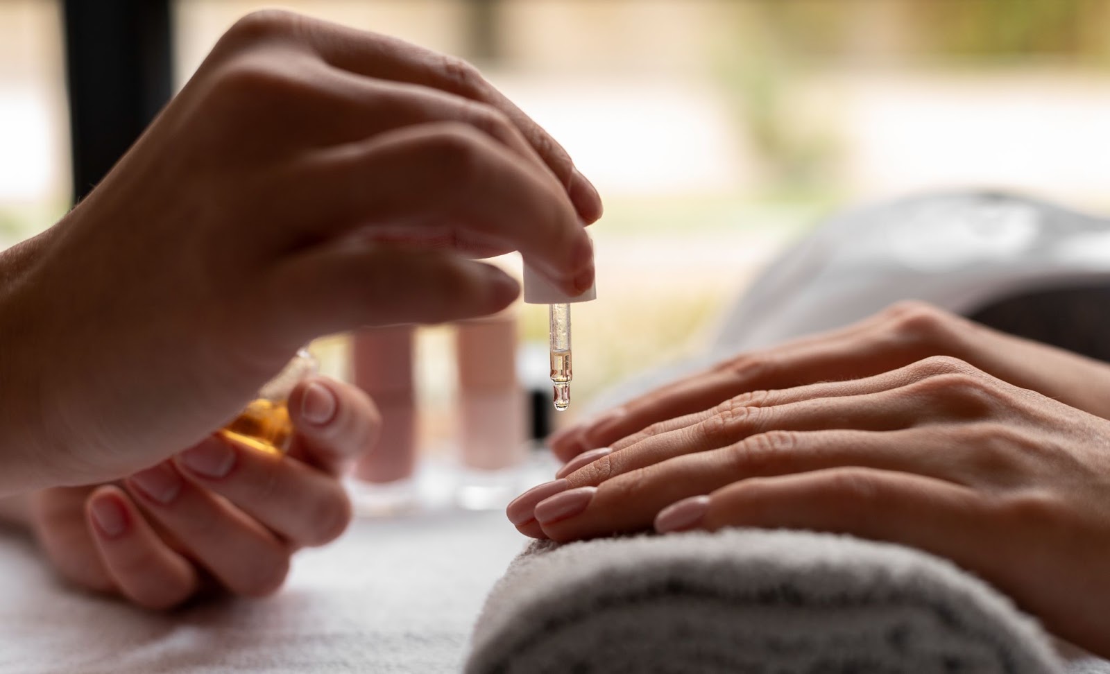 Na imagem há uma pessoa aplicando óleo nas unhas de outra pessoa, que apoia as mãos em uma toalha. A imagem representa a aplicação de óleo vegetal nas unhas para auxiliar na secagem.