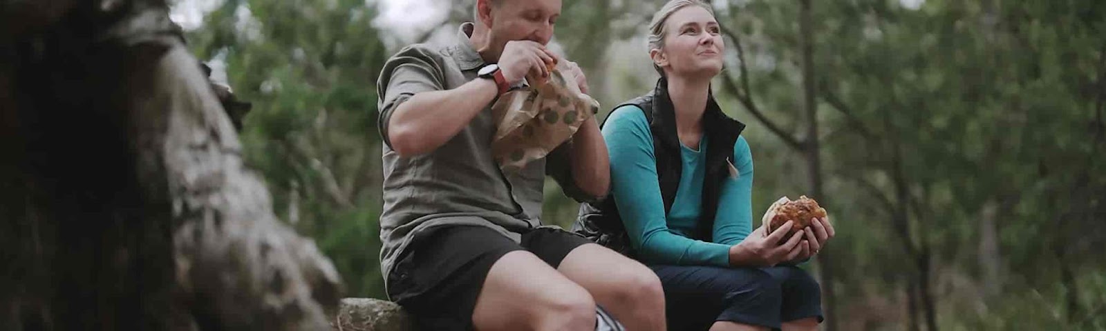 2 hikers sitting on a log on a nature trail, eating a Banjo's Bakery Salad Roll and smiling.