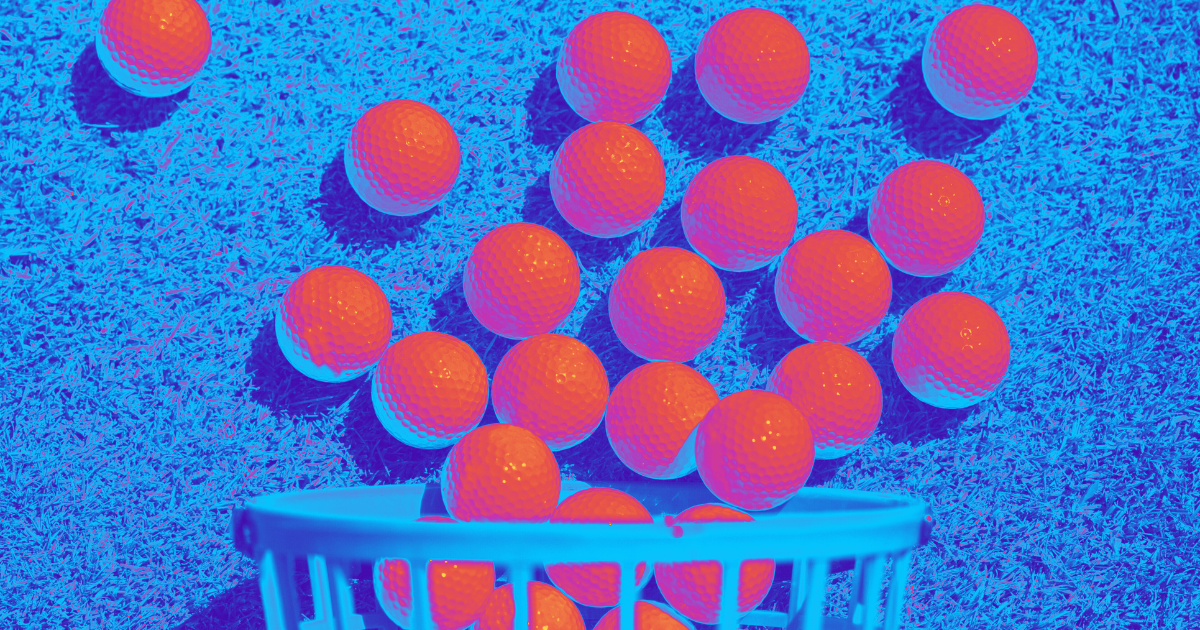 An orange golf ball basket stands on a grassy surface in blue hues, suggesting either an indoor driving range practice session or collection at an offsite driving range.