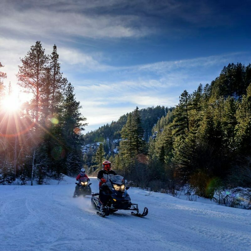 A picture depicting two snowmobile riders heading down a gradual slope at the Black Hills Snowmobile Trail in South Dakota, with evergreen trees and large hills in the background.