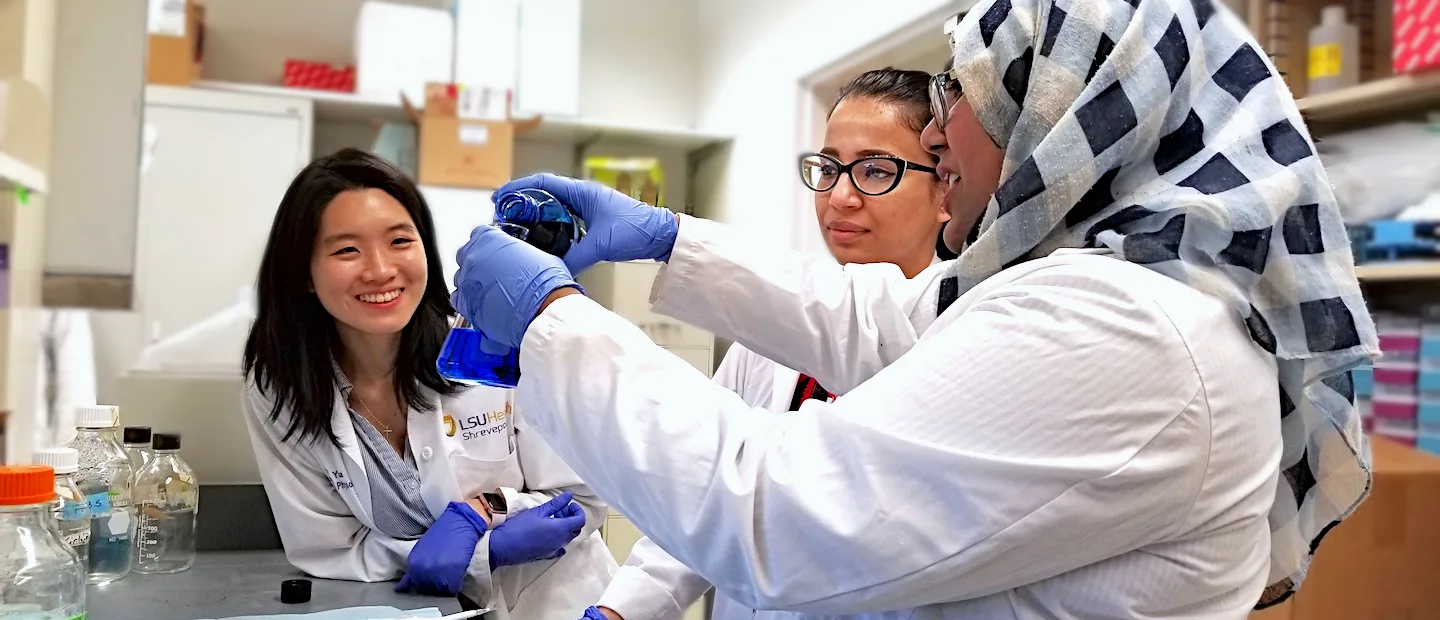 A photo of researchers in a laboratory at LSU Shreveport School of Medicine, featuring a person in a lab coat working with equipment in the foreground and shelves of scientific equipment and supplies in the background.