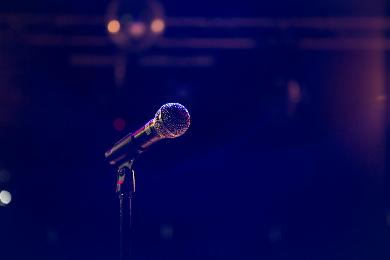 A microphone on a stand against a dark blue and pink bokeh background. Entrepreneur motivational speakers excel at customizing their messages to align with the specific goals and culture of an organization, a skill that turns a generic speech into a transformative experience for the audience.