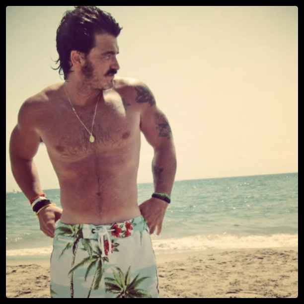 male with large side burns posing shirtless in floral board shorts at the beach in an instagram filter image 