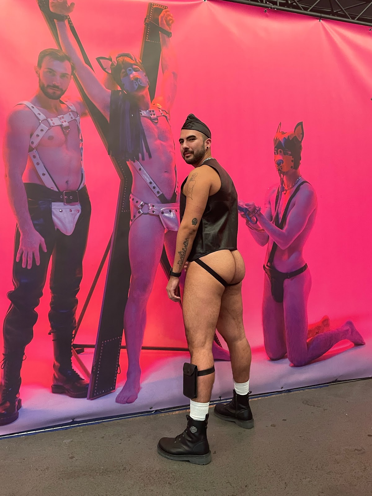 gay onlyfans content creator Phil posing in front of a darklands 2024 poster filled with puppy play models and gay males in harnesses wearing a leather vest and black jockstrap