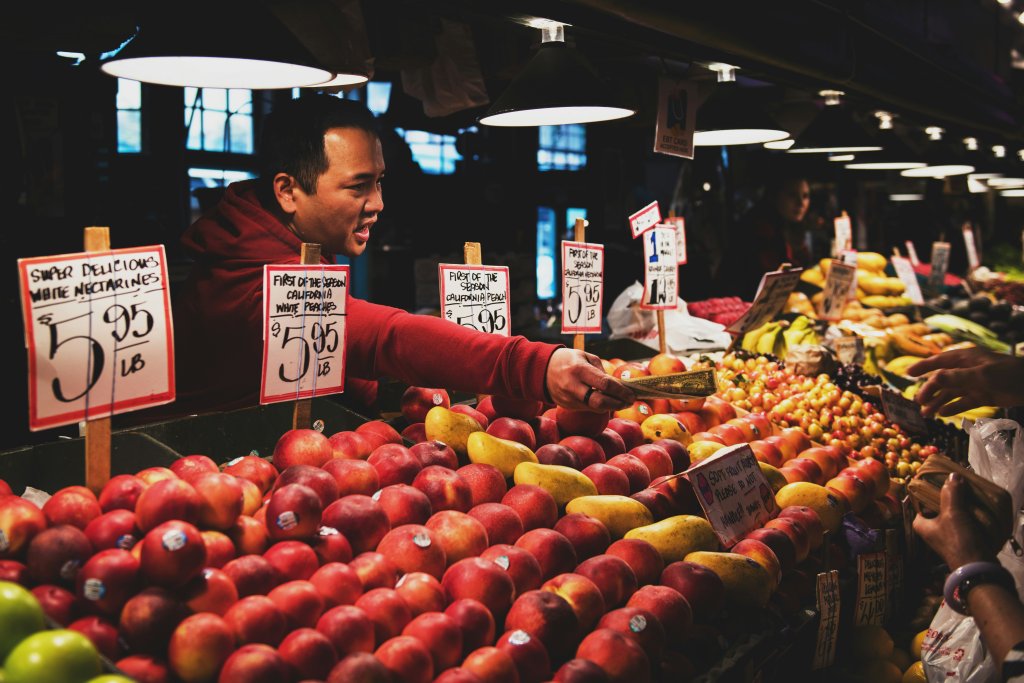 A imagem retrata uma cena em um mercado interno, onde várias frutas estão expostas em uma barraca. Entre as frutas visíveis, estão maçãs, bananas e limões. Alguns preços por quilo estão indicados em etiquetas. O braço de uma pessoa está estendido em direção às frutas, possivelmente selecionando ou apontando para elas.