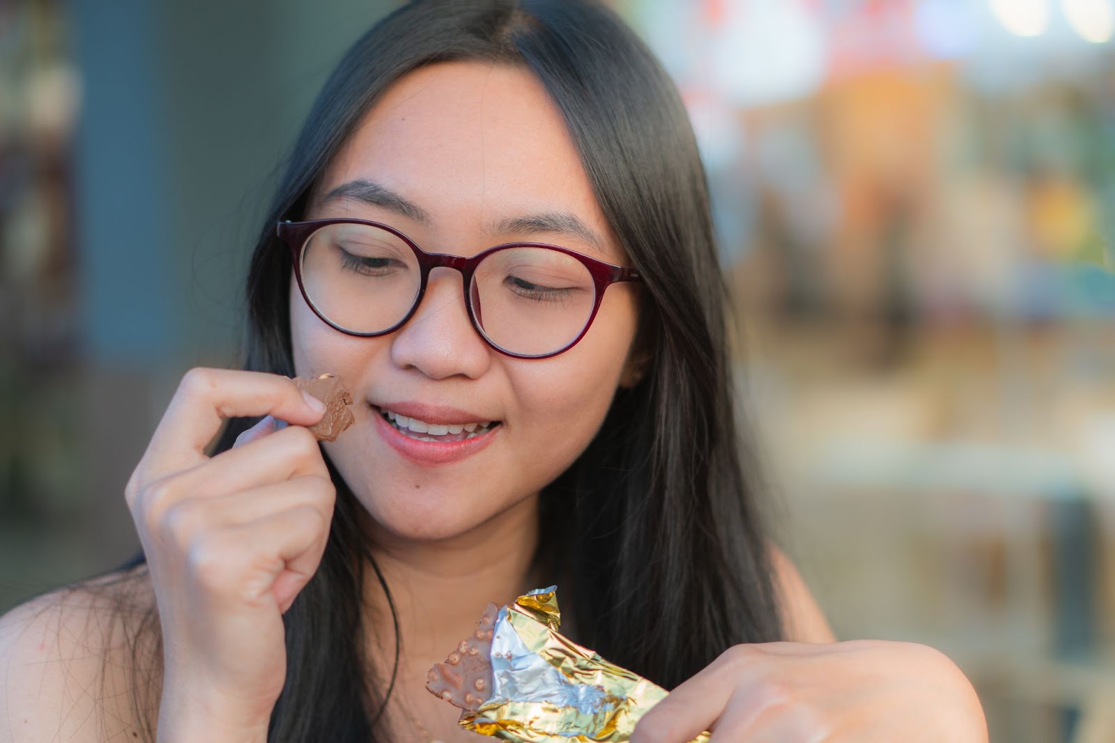 Girl with glasses eating a chocolate bar in a wrapper.