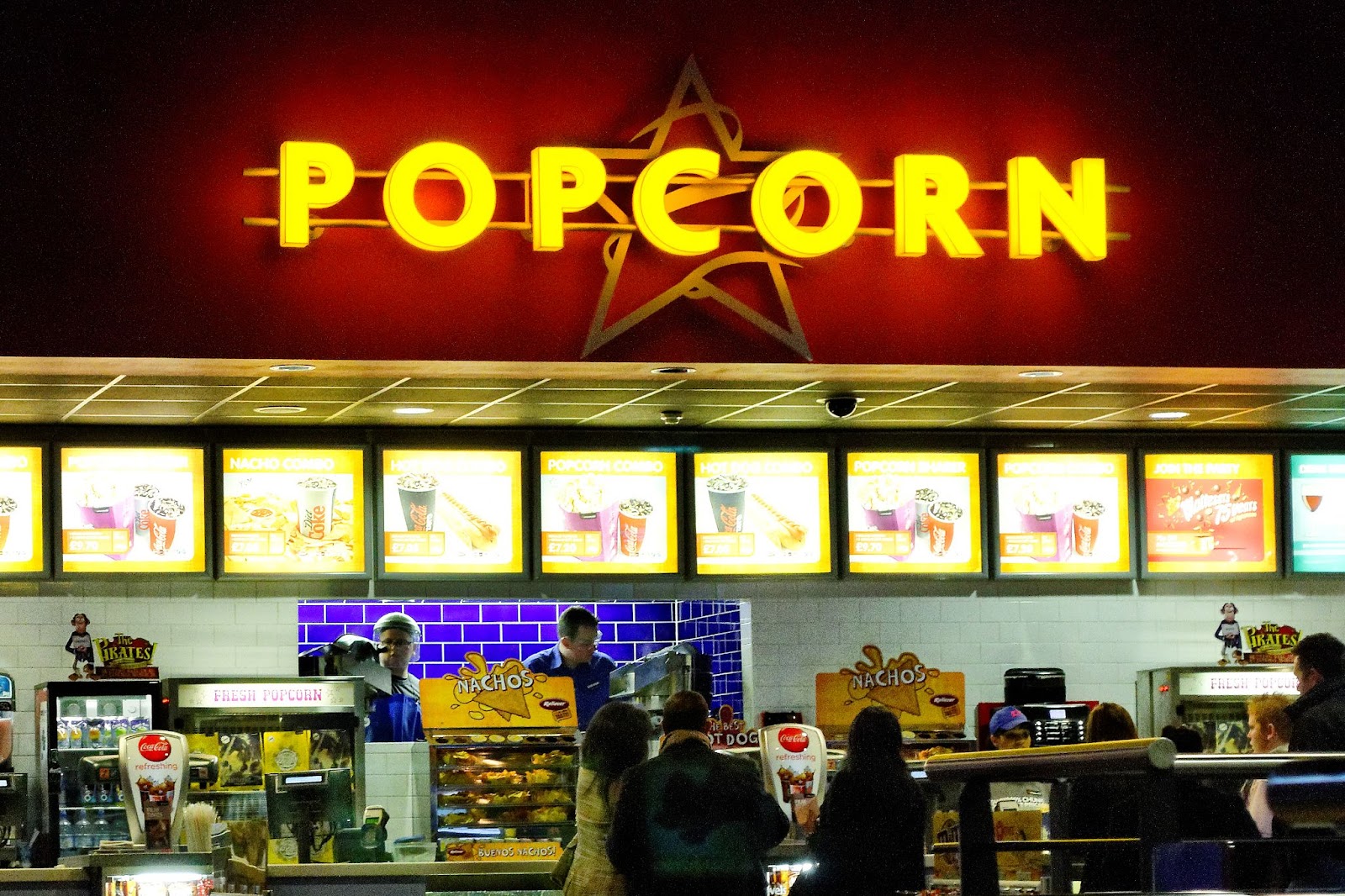 Bright golden 'POPCORN' letters on a red wall above movie theater concession stand; patrons lined up to buy snacks.