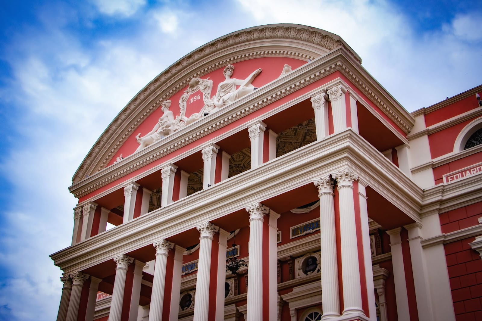 Parte da fachada frontal do Teatro Amazonas. Ela é pintada em cor-de-rosa, com colunas e detalhes esculpidos em branco, no estilo neoclássico.