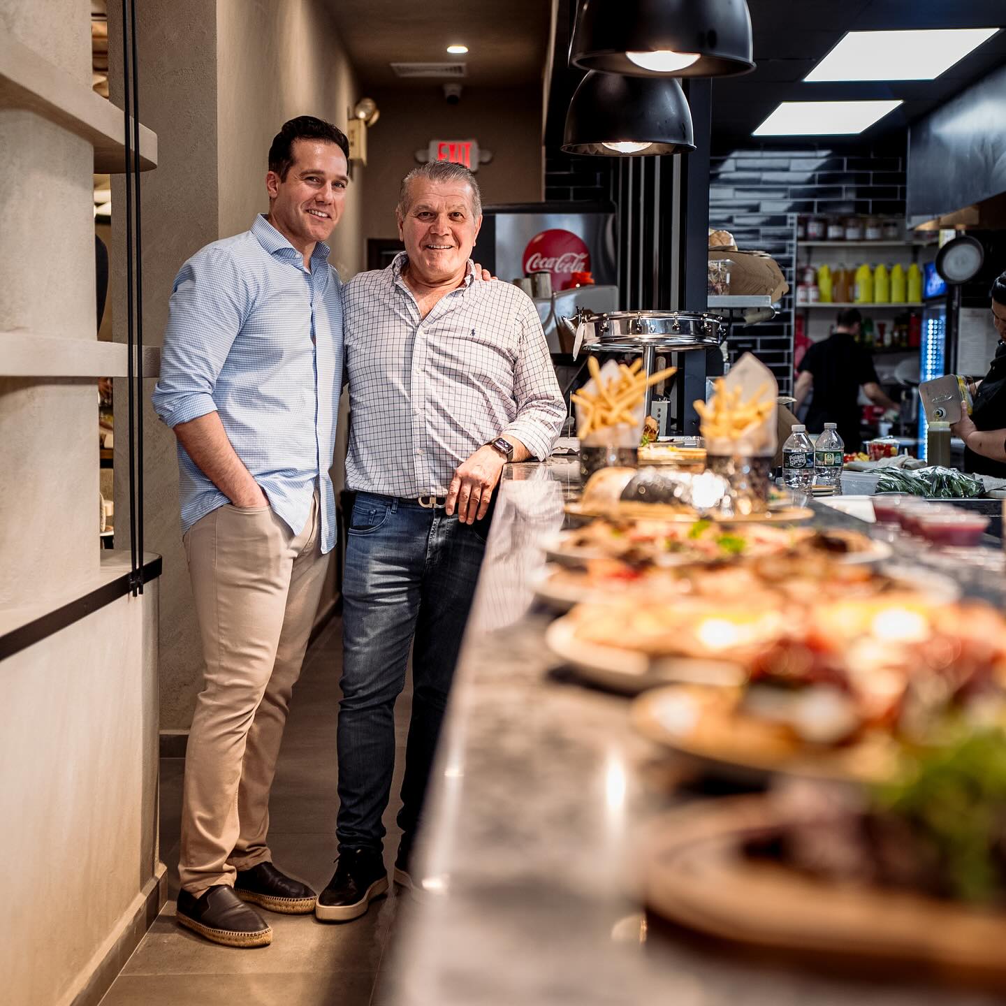 Kosta and Christos Nikolos smile in the kitchen at Carmine's in front of plates of food