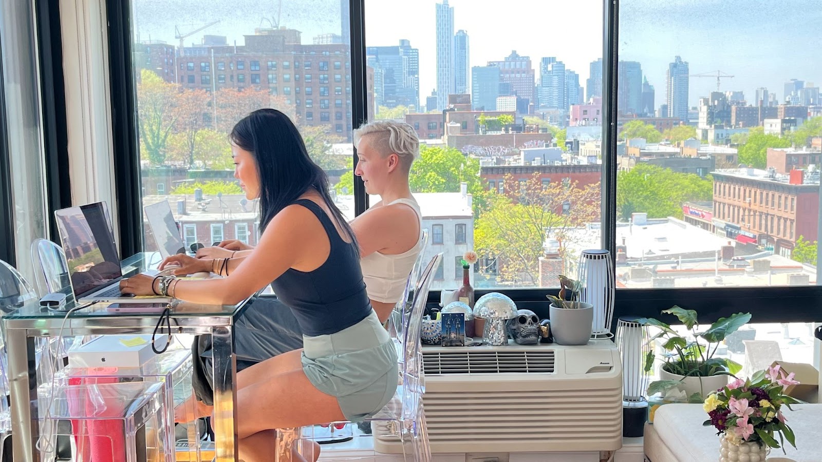 A beautiful Asian women with long black hair, in green running shorts and black tank top, works at her laptop at a high top table alongside Tarzan who is also staring at her laptop - a panoramic view of Brooklyn is all them, new leaves budding everywhere on the trees below. They are on the fourth or fifth floor.