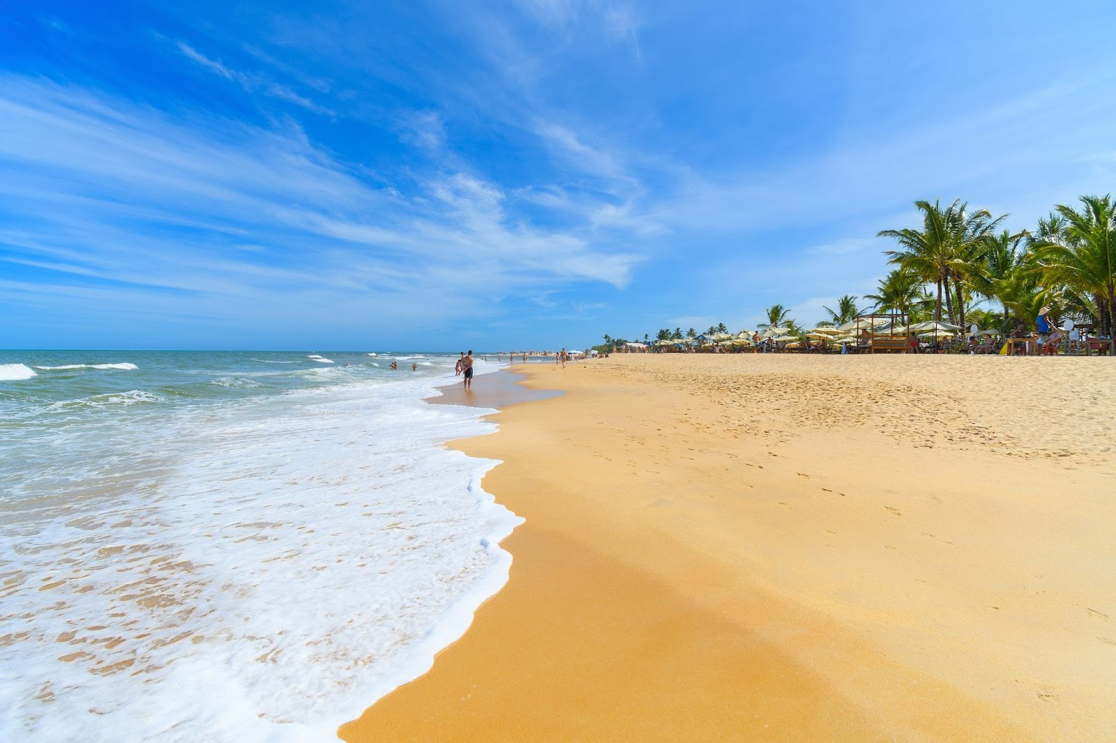 Larga faixa de areia da Praia do Mucugê, com a espuma da água do mar azul ao lado esquerdo da imagem.