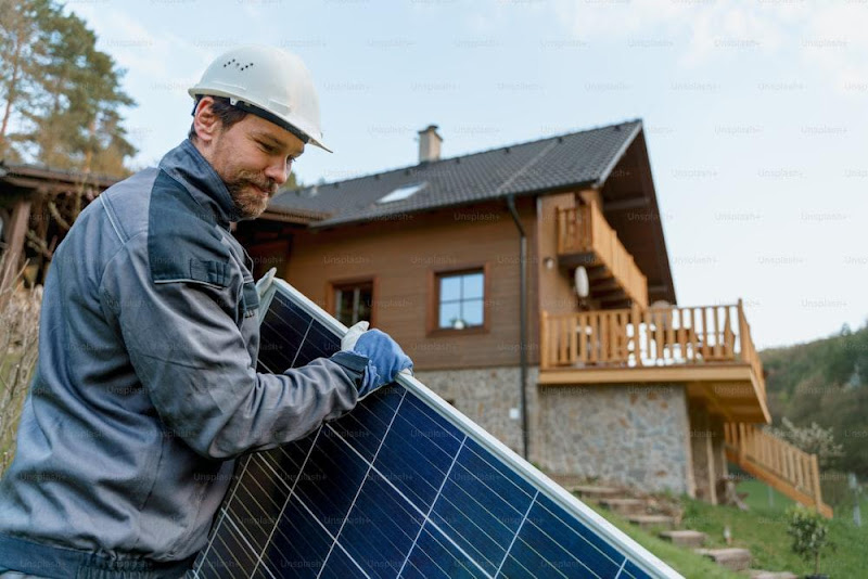 A smiling handyman solar installer carrying solar module while installing solar panel system on house.