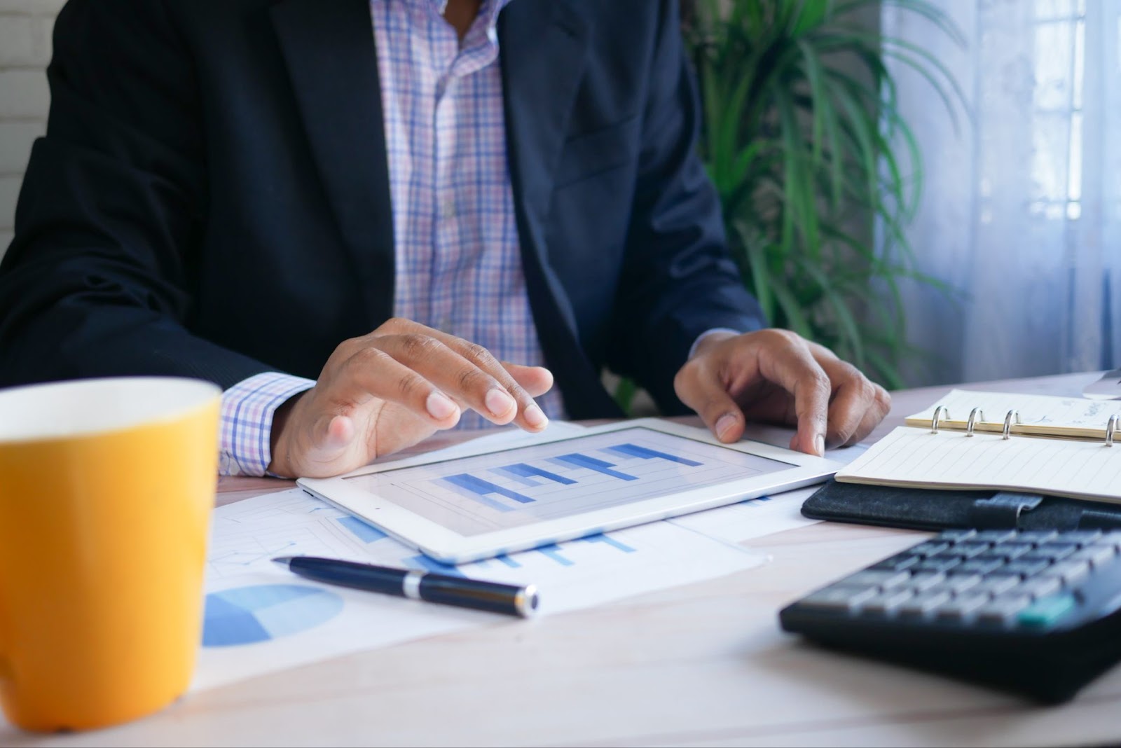 A man sat on his office desk. He is wearing a blue blazer and a plaid shirt. There is a cup of coffee, a pen, a calculator, a binder notebook, and a tablet on his desk. He is tapping on the tablet that displays four charts in the blue color. 