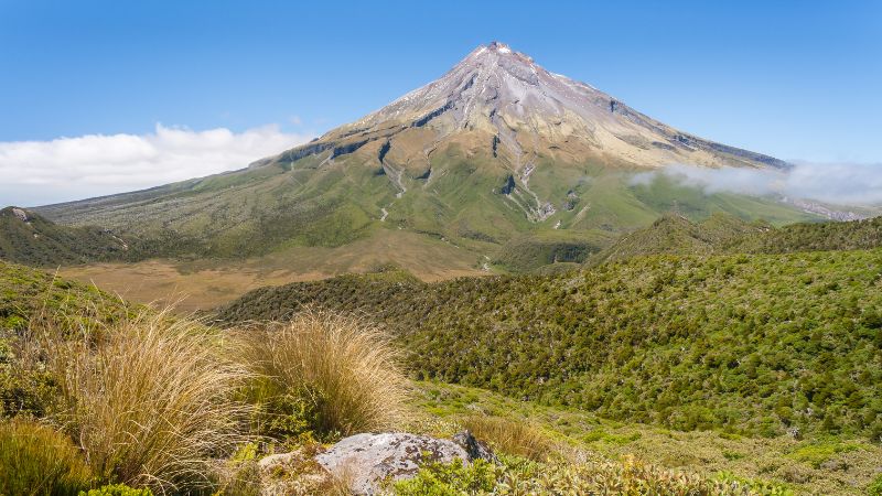 Lush vegetation surrounds the prominent volcanic cone of Mount Taranaki in Egmont National Park, under a clear blue sky.