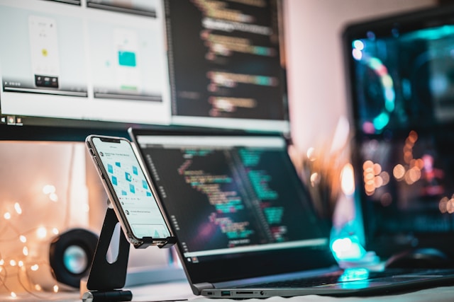 A desk with a phone laptop and a monitor showcasing UI/UX design