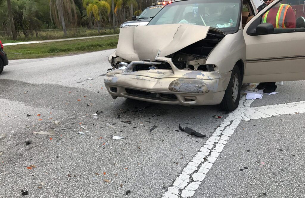A tan car with a smashed front end from a car accident sits in the middle of the road surrounded by broken glass and debris from the accident in spring hill, FL.