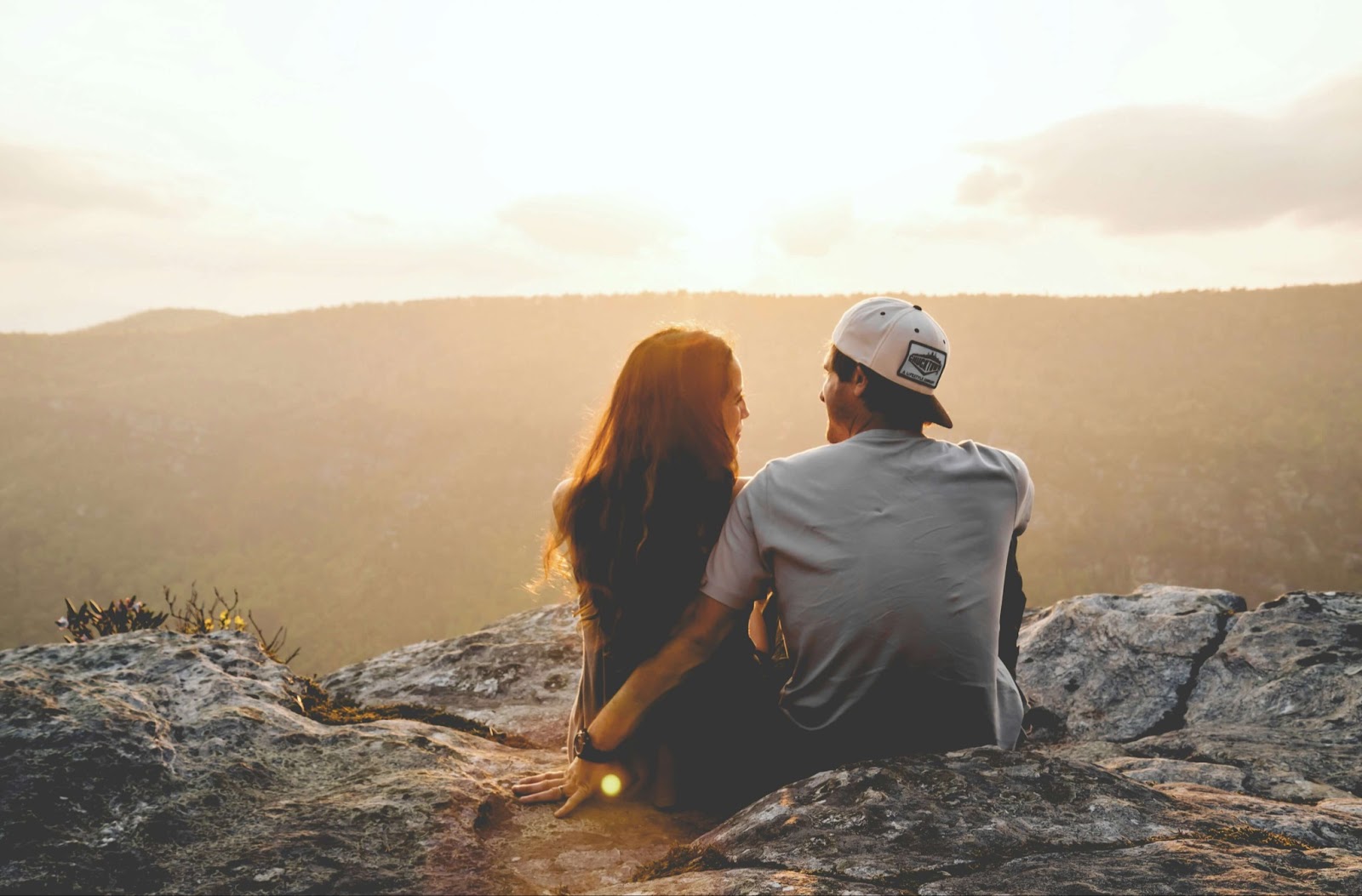 man and woman sitting on rock looking at each other with a view of mountain and sunset in the distance.