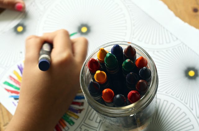 A child uses a crayon from a nearby jar of crayons to color a piece of paper