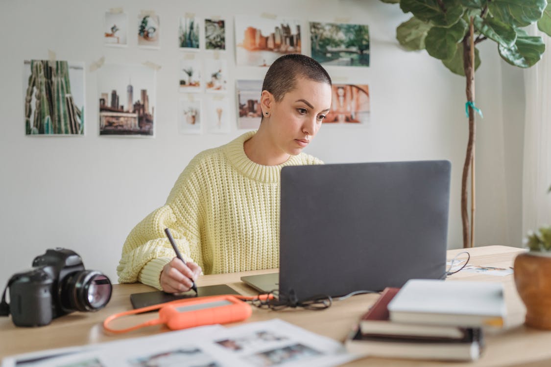 Free Thoughtful female in casual sweater working on netbook and drawing on graphic tablet in light room Stock Photo